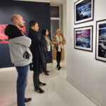 Zdjęcie z wernisażu wystawy iLand Małgorzaty Mikołajczyk. Dwóch mężczyzn i dwie kobiety rozmawiają i oglądają fotografie przedstawiające wzburzone morze lub ocean .
