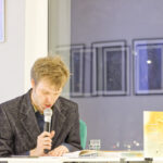 Zdjęcie z wieczoru poetyckiego Co kryje poezja obrazów Rafała Bakalarczyka. Artysta czyta swój wiersz mówiąc do mikrofonu.