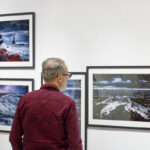 Zdjęcie z finisażu wystawy iLand Małgorzaty Mikołajczyk. Mężczyzna z brodą i w okularach ogląda fotografie przedstawiające falujący ocean.