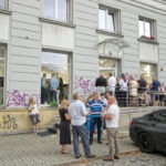 Przed wejściem do galerii. Goście stoją orzy drzwiach. Rozmawiają lub palą papierosy