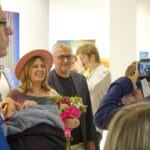 Wernisaż. Współautorka wystawa Joanna Pęgier. Pozuje do zdjęcia z bukietem w ręku. Jeden z gości robi jej zdjęcie telefonem