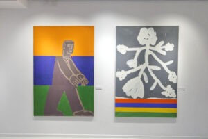 Ekspozycja wystawy "Paint and Print?". Na ścianie 2 obrazy. Pierwszy przedstawia człowieka na pomarańczowo-niebiesko-zielonym tle. Drugi przedstawia białą roślinę, a pod nią kolorowe pasy.