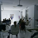 Spotkanie w Aptece Sztuki o nazwie "Sztuka Stand-upowania". Mężczyzna mówi do mikrofonu. Goście słuchają wystąpienia.