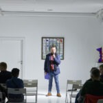 Spotkanie w Aptece Sztuki o nazwie "Sztuka Stand-upowania". Mężczyzna mówi do mikrofonu. Goście słuchają wystąpienia.